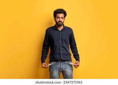 Poverty Concept. Portrait Of Upset Young Indian Man Showing His Empty Pockets And Looking At Camera, Broke Poor Millennial Guy Posing Over Yellow Background, Suffering Financial Crisis, Copy Space - Powered by Shutterstock