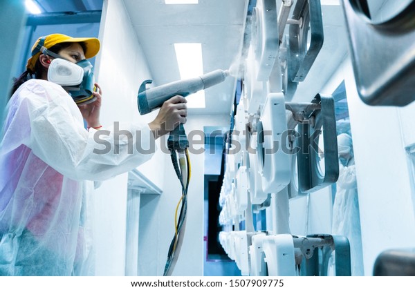 Powder coating\
of metal parts. A woman in a protective suit sprays white powder\
paint from a gun on metal\
products
