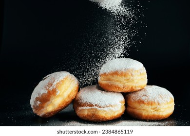 Powdered sugar is poured onto donuts. Traditional Jewish dessert Sufganiyot on black background. Celebrating Judaism holiday. Cooking fried Berliners.