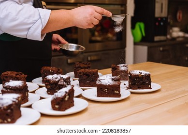 The Powdered Sugar Is Sprinkled Through A Sieve Onto Portions Of The Brownies. The Chef's Hand Holds The Sieve And Pours The Powdered Sugar Over The Desserts. Many Servings Of Brownies On Plates.