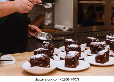 The Powdered Sugar Is Sprinkled Through A Sieve Onto Portions Of The Brownies. The Chef's Hand Holds The Sieve And Pours The Powdered Sugar Over The Desserts. Many Servings Of Brownies On Plates.