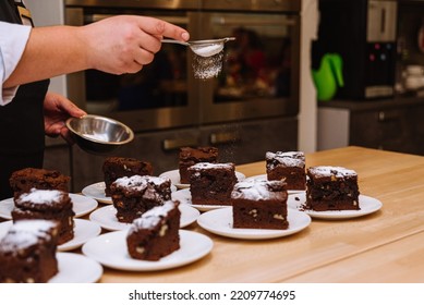 The Powdered Sugar Is Sprinkled Through A Sieve Onto Portions Of The Brownies. The Chef's Hand Holds The Sieve And Pours The Powdered Sugar Over The Desserts. Many Servings Of Brownies On Plates.