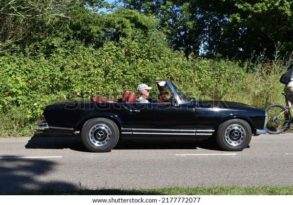 Powderham, Devon, UK. July 9th 2022.\
Mercedes SL classic car. 1960\'s convertible in black drives along a\
country lane. The driver wears a hat in the\
scene