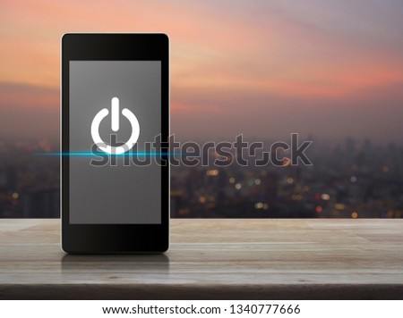 Power button flat icon on modern smart mobile phone screen on wooden table over blur of cityscape on warm light sundown, Start up business online concept