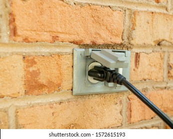 Power Cord Plugged Into Outdoor Electrical Wall Socket. Dual Covered Electricity Power Outlets On Brick Wall. Black Extension Cord Plug Inserted Into Exterior Wall Jack.