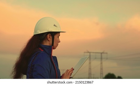 Power Engineer In A Protective Helmet Checks The Power Line, Digital Tablet. Civil Engineer, Woman Specializing In Electricity Supply, Working Outdoors. Green Energy Concept. Modern Technologies.