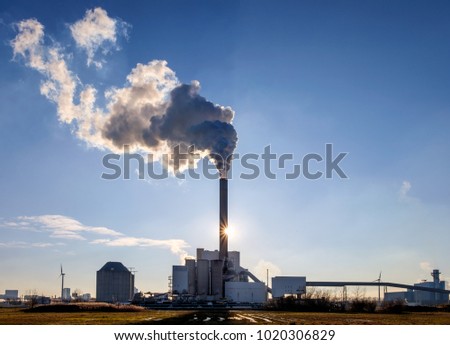 Power plant with a chimney and a large plume, shot against the light, with the sun just visible next to the chimney