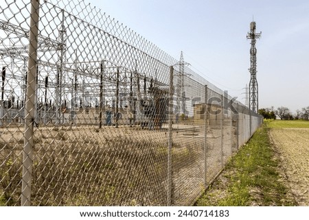 A power plant secured with electrical structures and a communications tower is visible behind a chain link fence on a clear day.