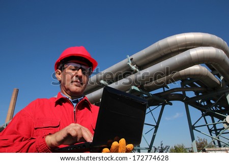Power Plant Worker With Notebook Computer. Industrial worker with notebook working in a power plant. An engineer in red overalls and hard hat against pipelines.