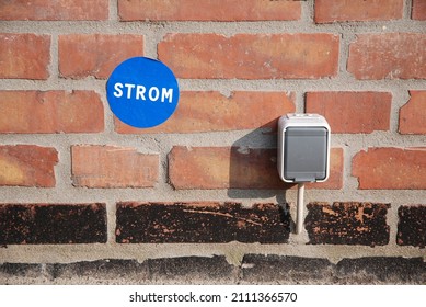 Power Socket Outdoor On A Brick House Wall With Blue Energy Sticker Written In German Strom - Engl. Electricity