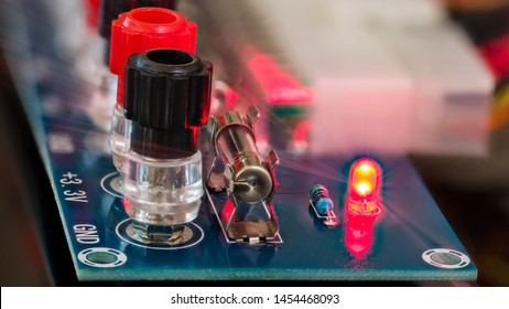 Power Supply Unit Diagnostics. Electronic Components Detail On Blue Circuit Board. Screw Terminals, Miniature Fuse, Shining Red LED Diode With Light Rays, Resistor And Blurred Connector In Background.