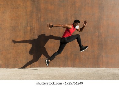 Powerful Athlete Training With N95 Face Mask For Protecting Against Covid-19. Young Black Man Running And Jumping Casting Shadow On A Wall.