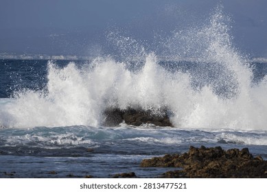 A powerful Atlantic wave violently explodes into a dramatic freeze-frame splash against dark shore reefs. The intense energy and raw strength of nature are highlighted against the deep blue sea and a distant coastline.