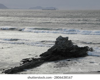 Powerful Atlantic waves breaking around a rocky outcrop with distant cargo ship and surfers waiting offshore at sunset. Rugged rocky outcrop stands prominently near the shoreline with powerful waves crashing around it. In the distance, a large cargo ship navigates the Atlantic Ocean under a dimming sky. Two surfers are visible offshore, waiting for waves. The scene captures the dynamic interaction of ocean elements against a backdrop of distant, silhouetted landforms.