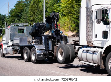 A Powerful Big Rig Semi Truck Tractor Tows A Broken White Semi Truck On A Highway With Green Trees To The Repair And Diagnostic Site In A Specialized Repair Shop With A Qualified Mechanics