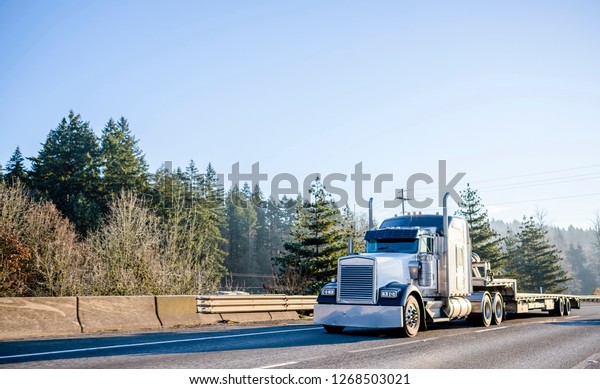 Powerful bonnet American idol black and white\
big rig long haul semi truck with empty step down semi trailer\
moving on wide highway with safety fence to warehouse for loading\
cargo for delivery