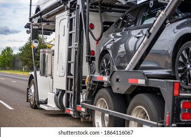 Powerful bonnet Big rig white car hauler semi truck tractor transporting different cars on two level adjustable hydraulic semi trailer running on the divided highway road with green trees on the side