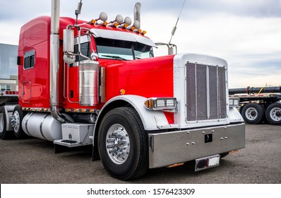 Powerful bright red classic big rig semi truck tractor with chrome accessories and signal horns on the roof and flat bed semi trailer standing on warehouse industrial parking lot waiting for load
