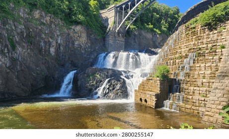 Powerful cascading waterfall flows over ancient stone dam structures beneath a large steel arched bridge on a bright summer day
