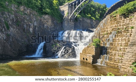 Powerful cascading waterfall flows over ancient stone dam structures beneath a large steel arched bridge on a bright summer day