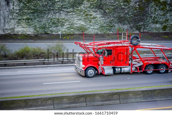 Powerful classic big rig bright red car hauler semi\
truck with eco - friendly diesel engine transporting empty two\
level semi trailer running on the divided road with ivy covered\
wall on the side