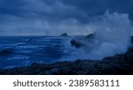 A powerful crashing wave can be seen crashing against the rocky shoreline, illuminated by a dark and ominous sky