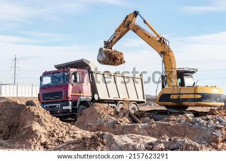 A powerful crawler excavator loads the earth into a dump truck against the blue sky. Development and removal of soil from the construction site