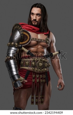 Powerful gladiator with a stylish beard wears lightweight intricate armor and poses gripping a feathered helmet against a grey background