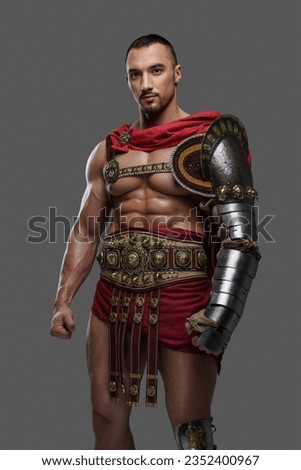 Powerful gladiator with a stylish beard wears lightweight intricate armor and poses gripping a feathered helmet against a grey background