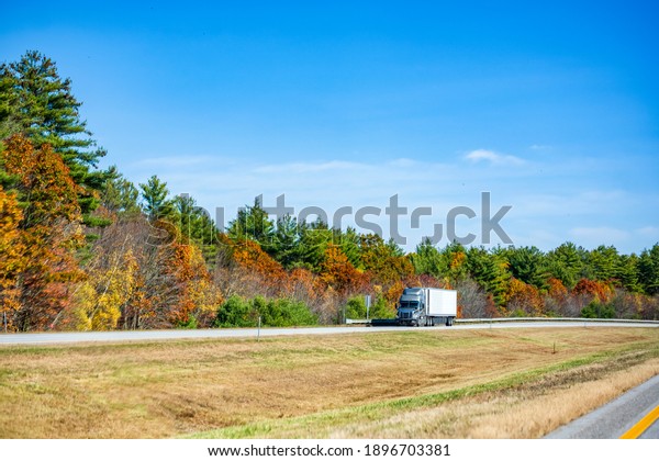 Powerful gray\
big rig industrial diesel semi truck with refrigerator semi trailer\
transporting cargo running on the divided highway road with\
colorful maples autumn trees in New\
England