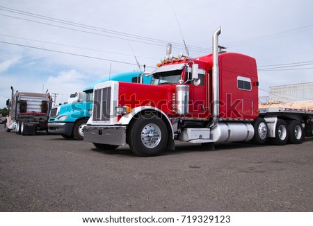 Powerful heavy-duty big rig red and blue classic and modern semi trucks with flat bed trailers  are parked in an industrial production park at the end of the working day