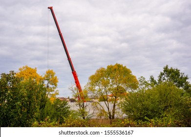 Powerful and huge telescopic arm of a mobile crane truck, hidden among leafy trees, lifting a load by means of cables to set up a prefabricated wall on a construction site