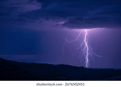 A powerful lightning bolt dramatically illuminates the night sky above a dark, silhouetted forest