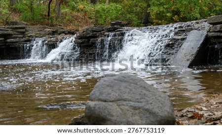 A powerful low-angle photograph capturing an RC truck perched on top of a rocky waterfall, surrounded by flowing water and rugged terrain. A mid-sized rock in the foreground adds depth and scale, enhancing the dramatic composition. This image highlights off-road RC adventure, motion, and natural rugged beauty.