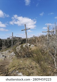 A powerful low-angle shot of three large, weathered wooden crosses standing prominently on a rugged, rocky hilltop under a dynamic blue sky filled with scattered white clouds. The central cross rises slightly higher than the two flanking it, closely resembling the traditional biblical imagery of Calvary or Golgotha. The crosses are constructed from rustic timber planks, showing signs of aging and exposure to the elements. The surrounding landscape features a sloping terrain covered in dry grasses, low-growing green shrubs, and wild bushes, with a dense coniferous and deciduous forest visible in the background valley. The natural daylight accentuates the spiritual, historical, and solemn atmosphere of the scene. This evocative image perfectly illustrates themes of Christian faith, Easter crucifixion, Good Friday, religious heritage, pilgrimage sites, and spiritual contemplation in a remote outdoor setting. Ideal for religious publications, historical documentaries, spiritual websites, Easter marketing materials, and conceptual designs focusing on devotion and sacred landscapes.