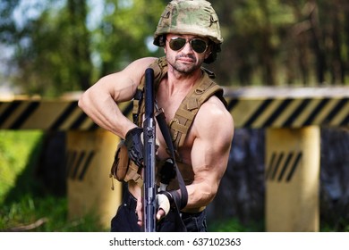 Powerful mercenary with submachine gun aiming the target near roadblock. Strong ripped man in helmet and sunglasses with rifle in hand. Soldier, mercenary, warrior, paintball, military concept - Shutterstock ID 637102363