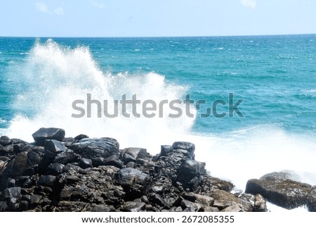 A powerful ocean wave crashes dramatically on large, dark rocks along a rugged coastline, creating a large splash of white foam under a clear blue sky