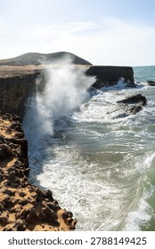 A powerful ocean wave explodes against steep rocky cliffs, sending dramatic sea spray high above the coastline under bright daylight. The swirling turquoise water, textured rocks, and intense surf capture the raw force and beauty of the coastal landscape.