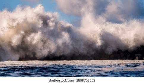 A powerful ocean wave explodes against a rocky shore, sending white foam and spray high into the air under a bright sky