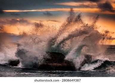 A powerful ocean wave explodes into a spray of water against a dark rock formation during a vibrant sunset, creating a dynamic natural spectacle