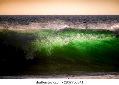A powerful ocean wave glows with emerald green light as it curls and breaks, illuminated by the setting sun