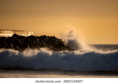 Powerful ocean waves are captured in motion, exploding against a rugged, dark rock formation under a warm, golden sky at dusk
