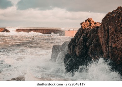 Powerful ocean waves crash dramatically against rugged dark rocks on a stormy day. Golden light illuminates the rocky coastline and a distant sea wall, creating a striking contrast with the turbulent sea and cloudy sky. This dynamic seascape evokes feelings of strength, resilience, and the raw beauty of nature.