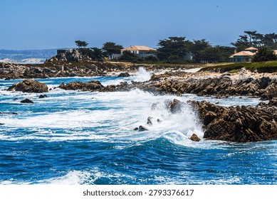 Powerful ocean waves crash dramatically against rugged rocky outcroppings along the scenic Monterey Peninsula coastline in California, USA. Turquoise and deep blue Pacific Ocean water surges and splashes into white sea foam as waves break over exposed rocks. Lush green cypress trees frame luxury coastal homes with terracotta rooftops perched along the shoreline in the background under a clear bright blue summer sky. This stunning coastal landscape captures the raw energy of the California coast, characteristic of the famous 17-Mile Drive area near Pebble Beach and Pacific Grove. Ideal for travel, nature, real estate, and lifestyle editorial and commercial use.