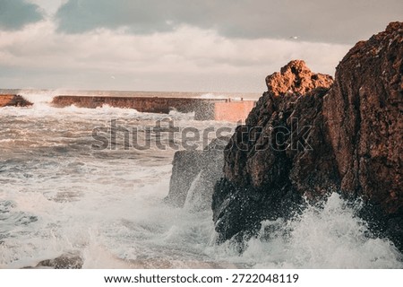 Powerful ocean waves crash dramatically against rugged dark rocks on a stormy day. Golden light illuminates the rocky coastline and a distant sea wall, creating a striking contrast with the turbulent sea and cloudy sky. This dynamic seascape evokes feelings of strength, resilience, and the raw beauty of nature.