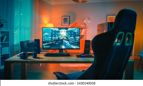 Powerful Personal Computer Gamer Rig With First-Person Shooter Game On Screen. Monitor Stands On The Table At Home. Cozy Room With Modern Design Is Lit With Warm And Neon Light.