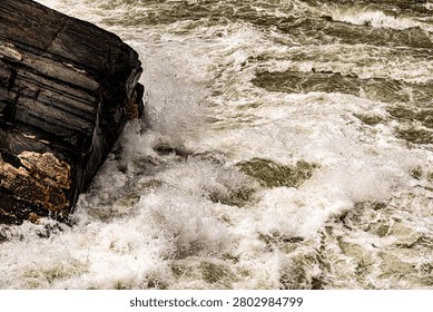 A powerful photograph capturing the interaction between a stationary, dark rock and the violent, rushing current of a river. The explosion of white spray and foam against the rock face emphasizes the untamed energy of the rapids.