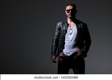 Powerful punk man posing with his hand in his pocket while wearing sunglasses, a black leather jacket and jeans, standing on black studio background