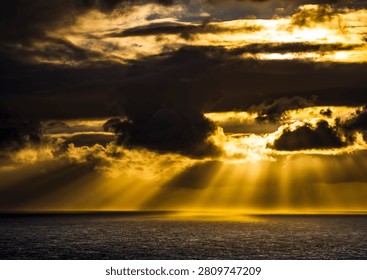 Powerful rays of sunlight pierce through heavy, dark clouds, illuminating the vast expanse of the sea with a golden glow