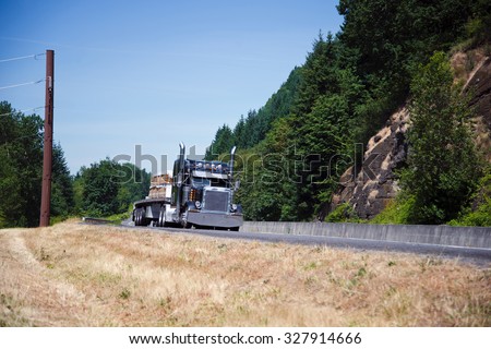 Powerful spectacular professional commercial classic dark blue semi - truck with an open trailer flat bed and chrome accents transports building boards on the highway near the picturesque cliffs.